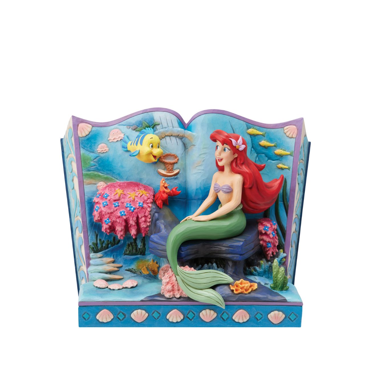 Introducing the Little Mermaid Storybook Figurine from Disney Traditions by Jim Shore. This piece tells the classic tale of Ariel, Flounder, and Sebastian, bringing this beloved story to life in a stunning 3D scene. The figurine depicts Ariel, Flounder, and Sebastian under the sea, surrounded by colourful plant life and shells that add a magical touch to this exquisite work of art.