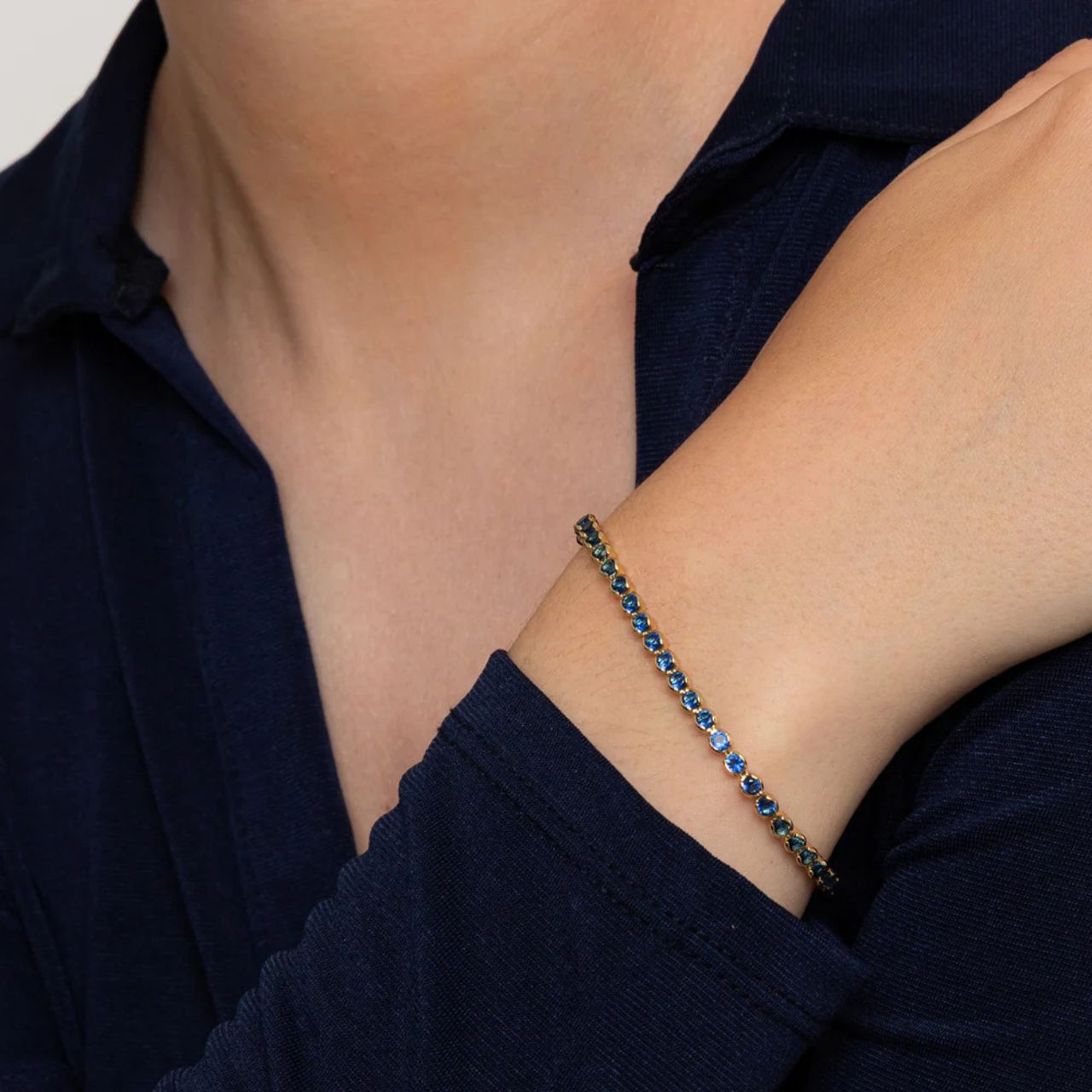 Miniature gold plated tennis bracelet with sapphire CZ stones. Elegant when worn alone and also ideal for layering with watch or other bracelets. Fold over clip fastening.