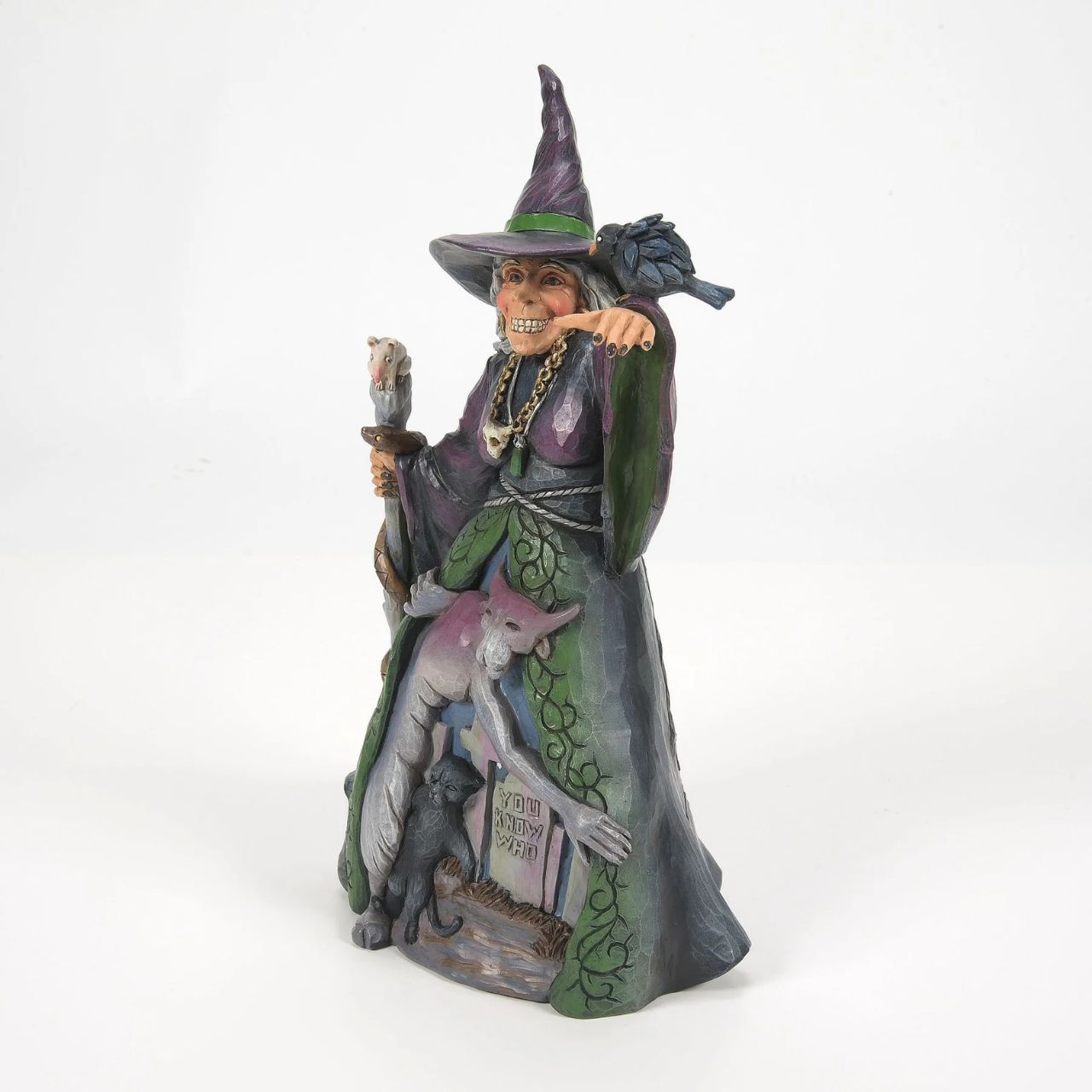 Enter the enchanting world of Halloween with this haunting Jim Shore witch figurine, adorned in ghastly purple and green robes, a perched crow and a staff with a slithering snake. Her cloak reveals a chilling graveyard scene with a spook and a black cat.