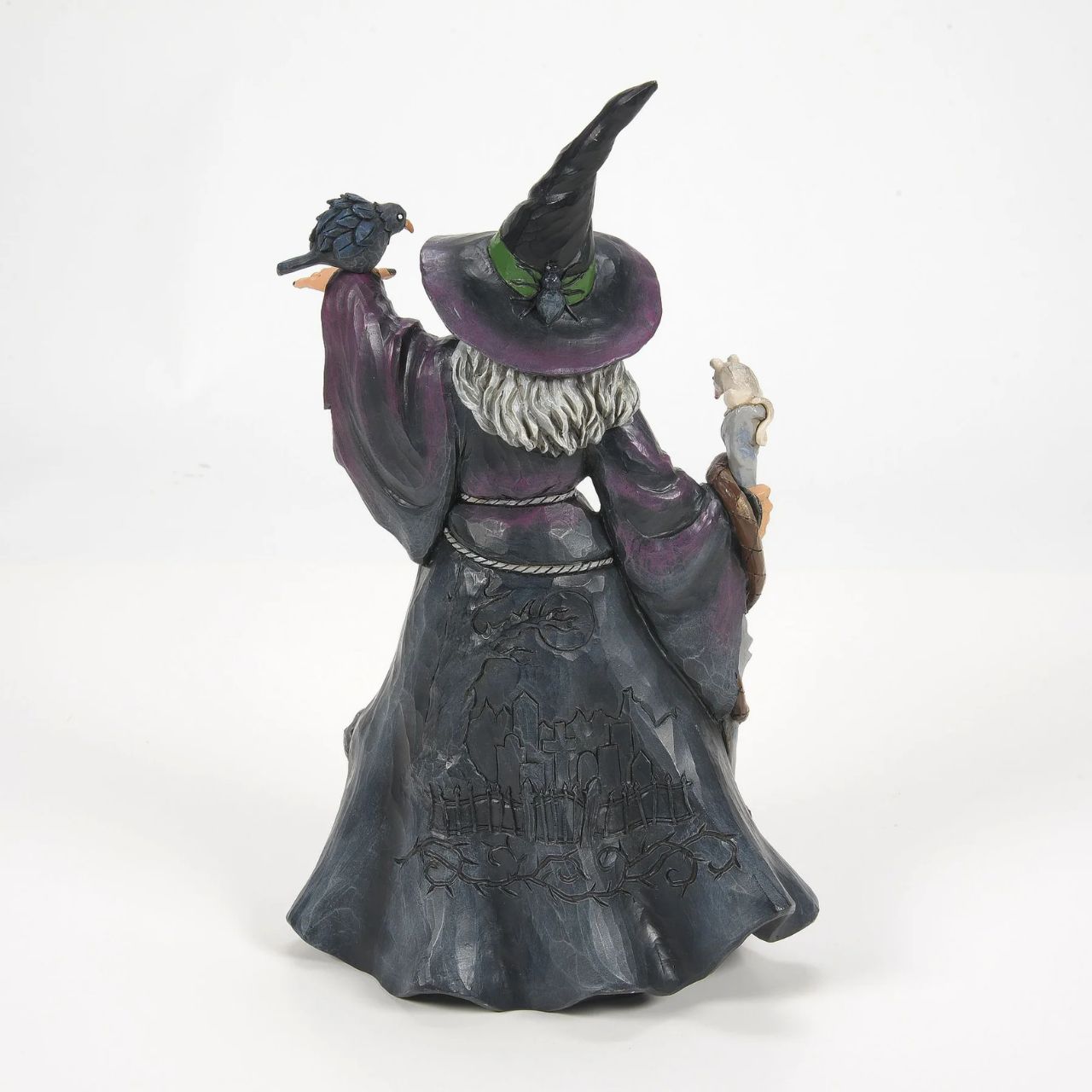 Enter the enchanting world of Halloween with this haunting Jim Shore witch figurine, adorned in ghastly purple and green robes, a perched crow and a staff with a slithering snake. Her cloak reveals a chilling graveyard scene with a spook and a black cat.