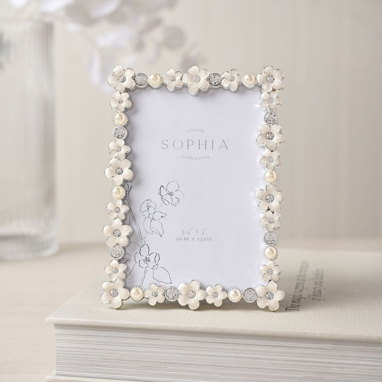 Introduce a touch of elegance and charm to your photo collection with the Sophia Classic Daisy 3.5” x 5” Frame in White. Featuring an intricate floral pattern and decorative crystals, this frame transforms your cherished memories into a stunning display piece that exudes sophistication.