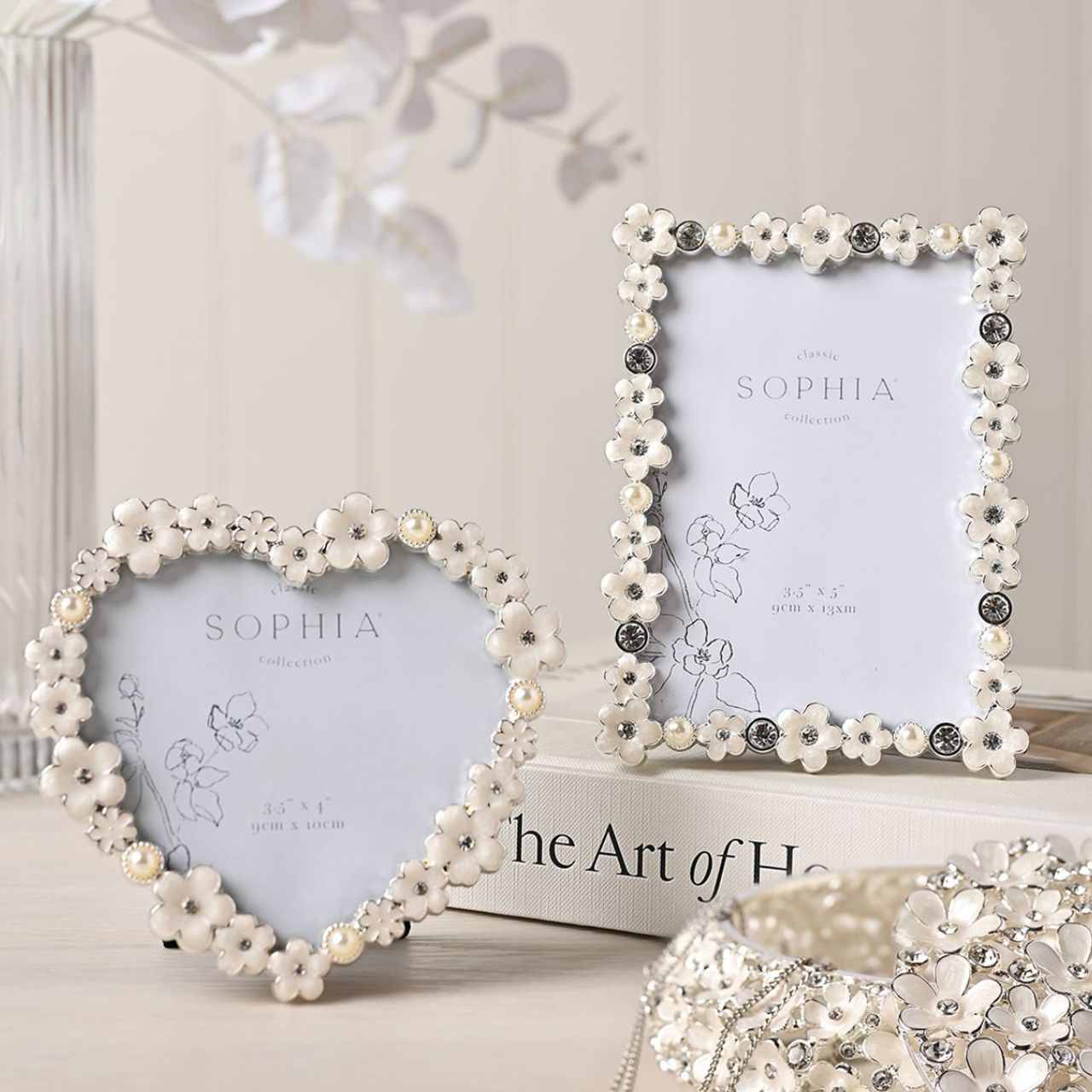 Introduce a touch of elegance and charm to your photo collection with the Sophia Classic Daisy 3.5” x 5” Frame in White. Featuring an intricate floral pattern and decorative crystals, this frame transforms your cherished memories into a stunning display piece that exudes sophistication.