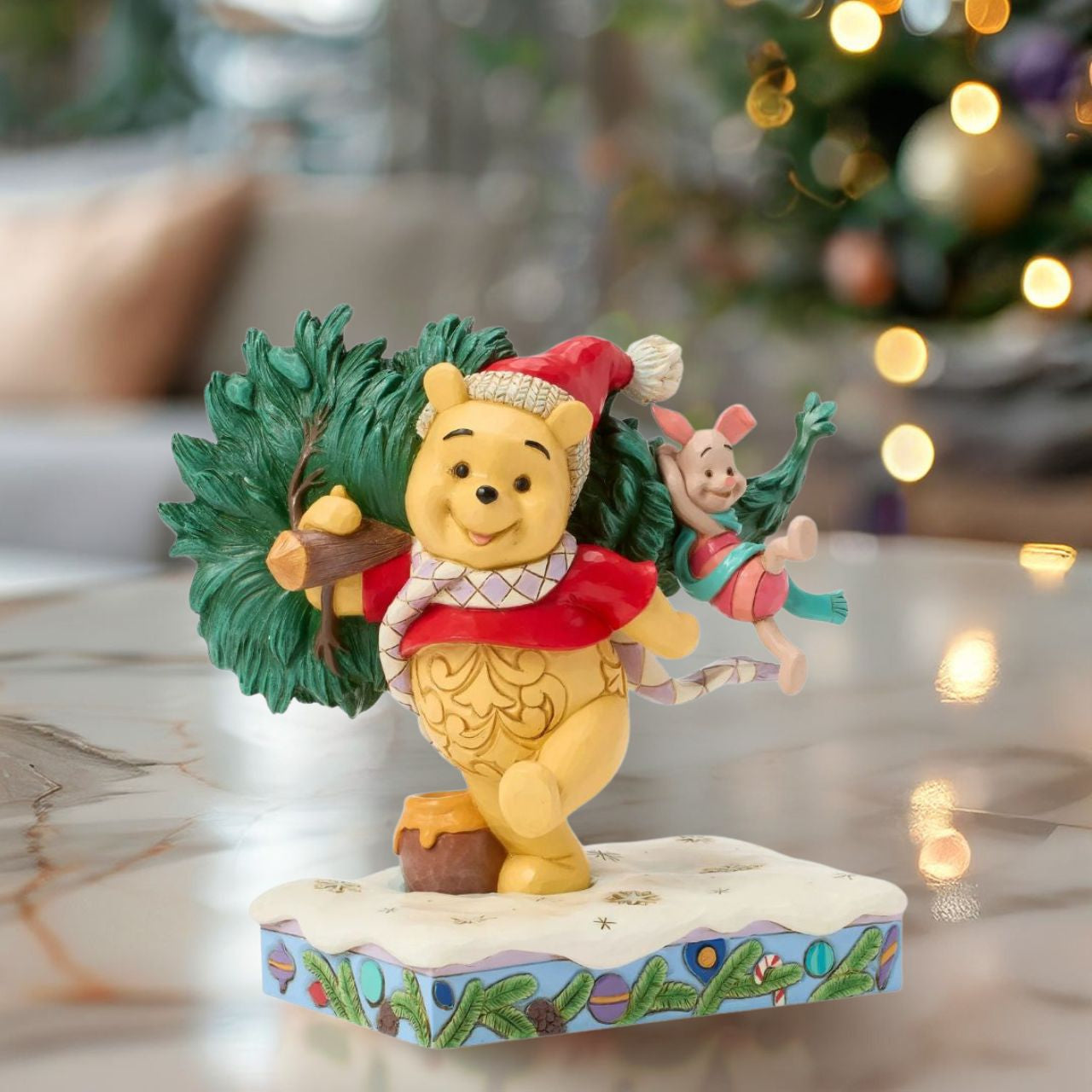 In this delightful Jim Shore scene, Winnie the Pooh sports intricate rosemaling and a Santa hat adorned with holly as he joyfully carries home a Christmas tree with Piglet in tow. They march through the snow on a festive stage of ornaments.