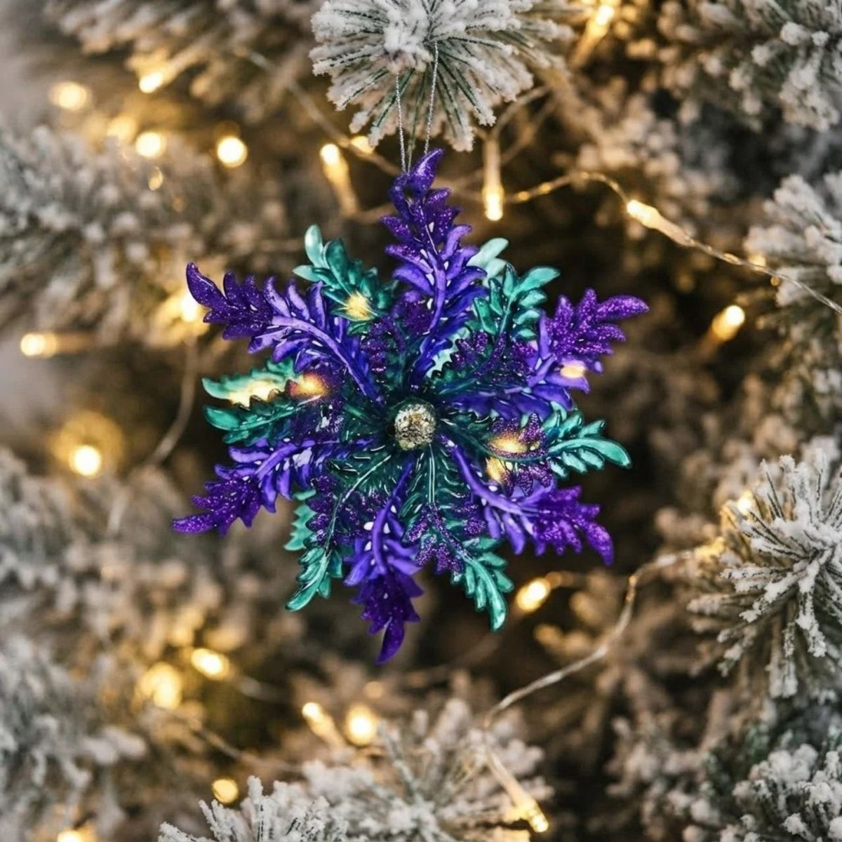 A glittery snowflake Christmas ornament in purple and teal hanging on a flocked Christmas tree with warm string lights.