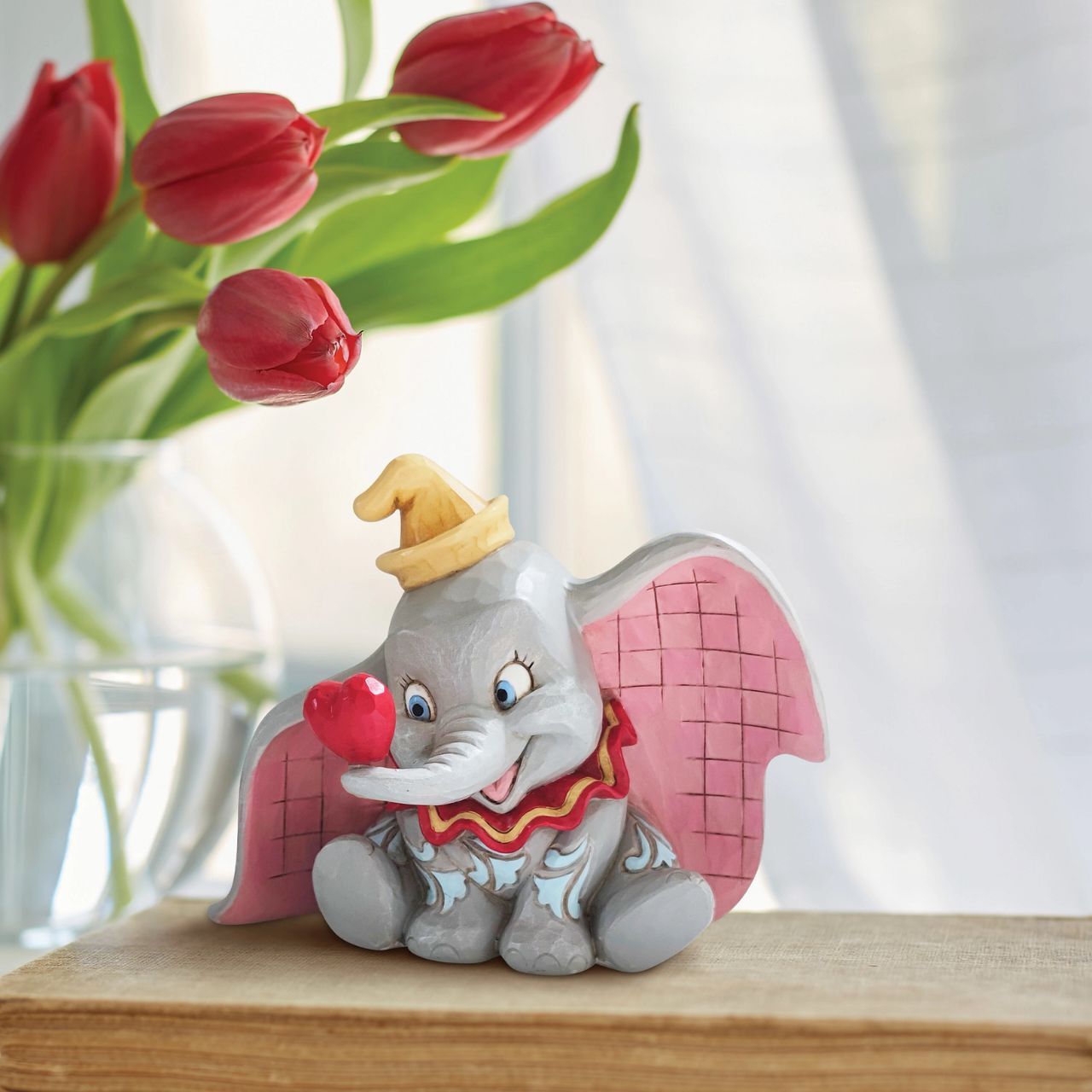 Disney Traditions A Gift of Love Dumbo with Heart Figurine  "A Gift of Love" Holding a heart with his trunk, the lovable flying elephant, Dumbo, smiles sweetly in this charming creation by Jim Shore. With pretty patchwork on his ears and rosemaled design on his skin, Dumbo looks dashing in this simple and sentimental design.