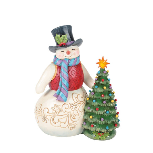 This enchanting Jim Shore snowman, dressed in a gorgeous tunic, scarf, and holly-brimmed top hat, stands beside a decorated Christmas tree and the star lights up. Adorned with snowflakes and rosemaling, he brings a vintage warmth to your holiday home.