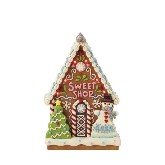This enchanting Jim Shore LED light-up sweet shop invites you into a festive gingerbread house with colourful icing, cookies, and candy. Glowing warmly, it promises a world of delicious treats and holiday cheer, making it a delightful centerpiece.