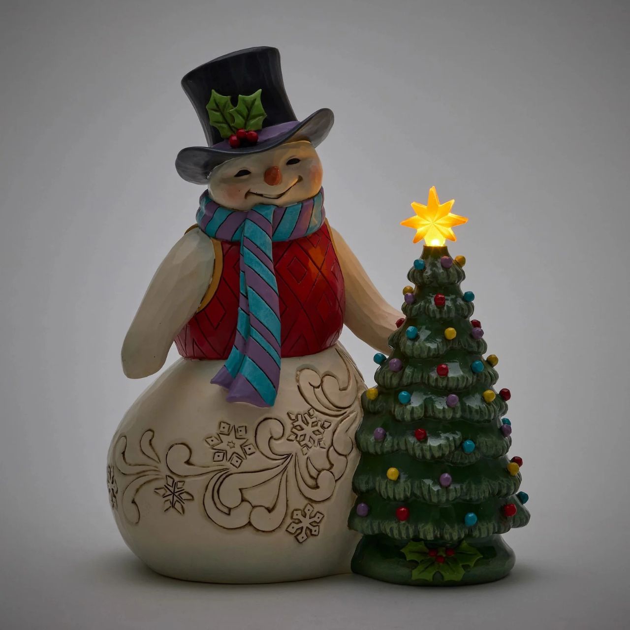 This enchanting Jim Shore snowman, dressed in a gorgeous tunic, scarf, and holly-brimmed top hat, stands beside a decorated Christmas tree and the star lights up. Adorned with snowflakes and rosemaling, he brings a vintage warmth to your holiday home.