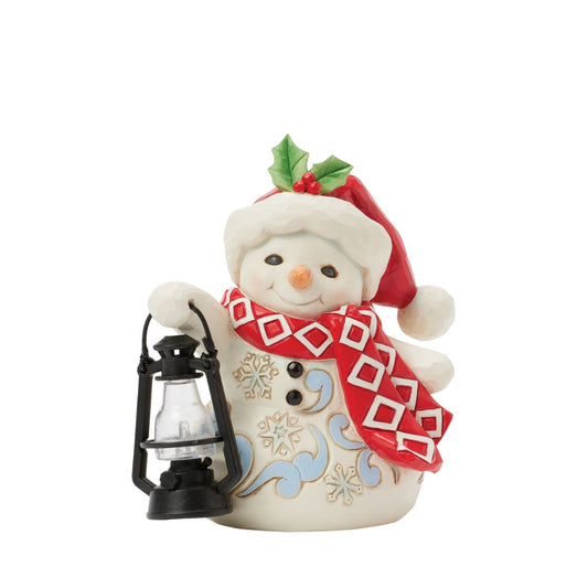 This delightful Jim Shore pint snowman carries a glowing candlelit lantern, spreading warmth and light. Dressed in a Santa hat adorned with holly, a patterned scarf, and snowflake accents, his rosy cheeks and bright smile spread Christmas cheer.