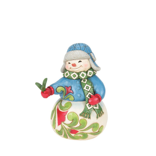 This sweet Jim Shore snowman, adorned in a patterned aviator hat and matching scarf, smiles warmly as he shares a picked pine sprig with you. His body and tunic feature delightful floral designs, adding a touch of charm to your winter decor.
