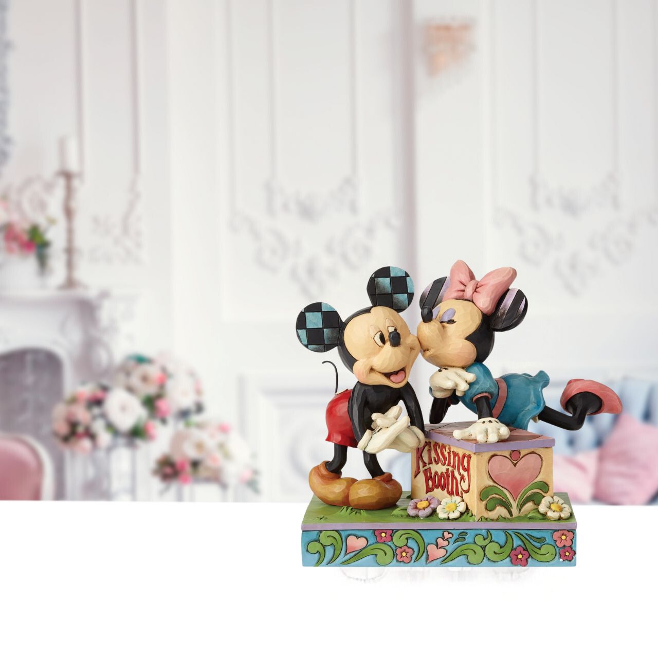 Minnie leans over a kissing booth to plant a smooch on one very smitten Mickey in this cheeky scene by Jim Shore. Handcrafted in his unique folk art style, the cast stone design features whimsical rosemaling and quilt patterns.