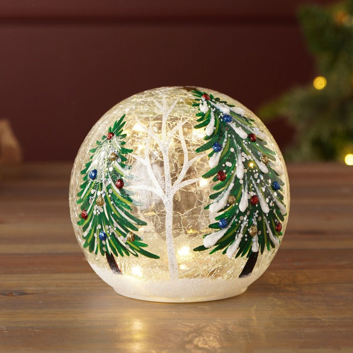 The LED Crackle Glass Light with Christmas Tree Design (Includes Try Me Button) adds a playful touch to classic Christmas lighting. Featuring a snowy winter scene with a bare tree and snow-covered evergreens adorned with ornaments, it glows softly, highlighting the crackle glass texture and festive details.