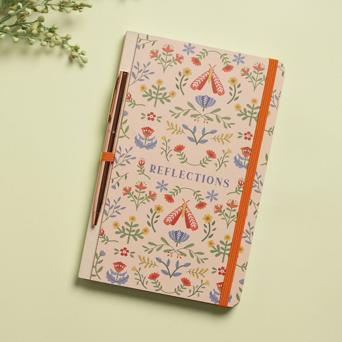 A floral-patterned notebook titled "Reflections" with an orange elastic closure pairs beautifully with a sleek pen, creating a set that's both useful and display-worthy. Jot down notes, memories, and dreams on smooth lined pages.