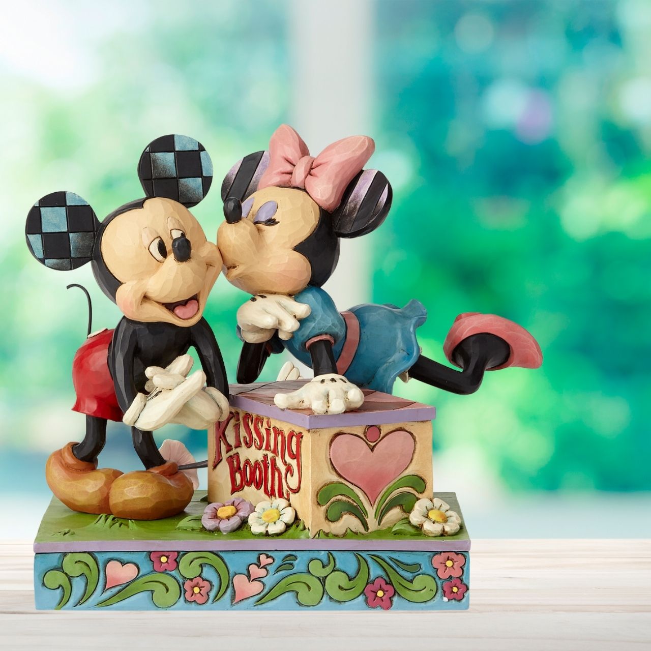 Jim Shore Kissing Booth Mickey Mouse and Minnie Mouse  Minnie leans over a kissing booth to plant a smooch on one very smitten Mickey in this cheeky scene by Jim Shore. Handcrafted in his unique folk art style, the cast stone design features whimsical rosemaling and quilt patterns. 