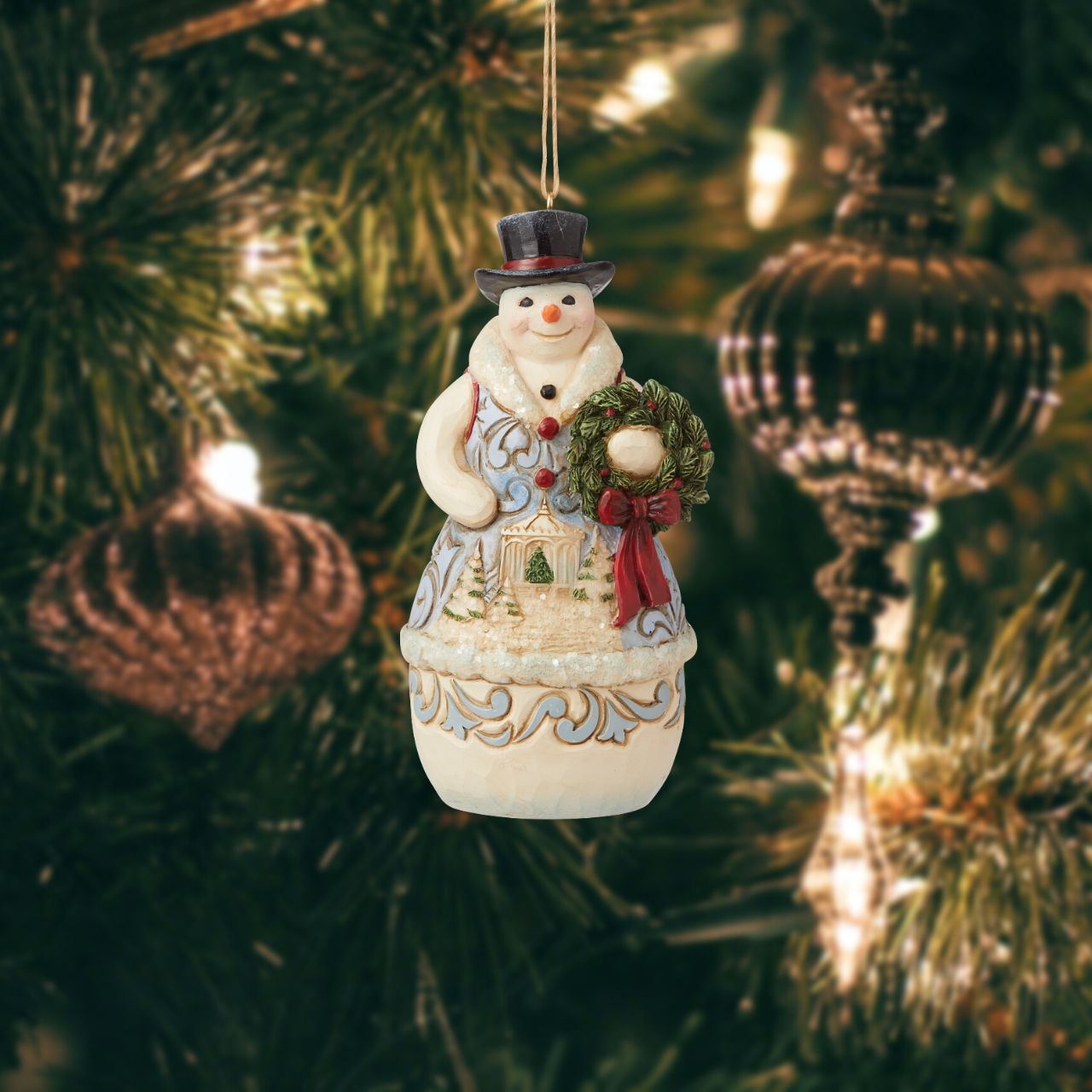 Jim Shore Heartwood Creek Victorian Christmas Snowman with Wreath Hanging Ornament  This Jim Shore Victorian snowman holds a wreath while a timeless winter scene plays across his chest. A smile on his face and a top hat on his head, he is ready for whatever the season brings. This snowman is brimmed in holly and holiday wonder.